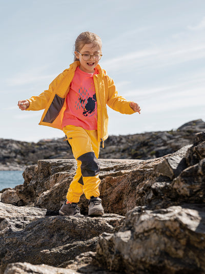 Didriksonsin lasten Briska hybriditakki keltaisessa sävyssä lapsen päällä ulkona kuvattuna. Lapsi kävelee kalliolla ja kuva on otettu edesä. 