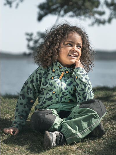 Lapsi istuu nurmikolla, ja hänellä on päällään Didriksonsin lasten vihreät Dusk-ulkoiluhousut