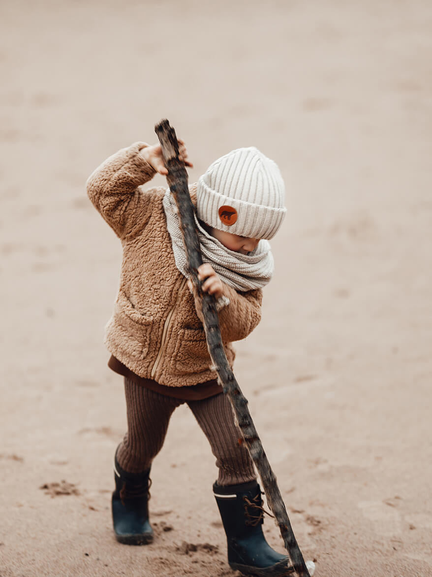 Lapsella on vaniljan värinen Superyellow Björn merinovillapipo, ja hän leikkii isolla kepillä rannalla.